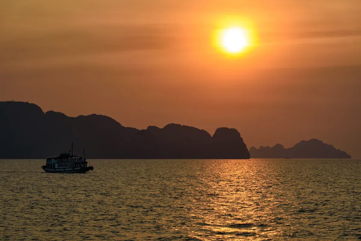 Sun setting behind karst silhouettes with a boat on the water, Ha Long Bay junk boat cruise